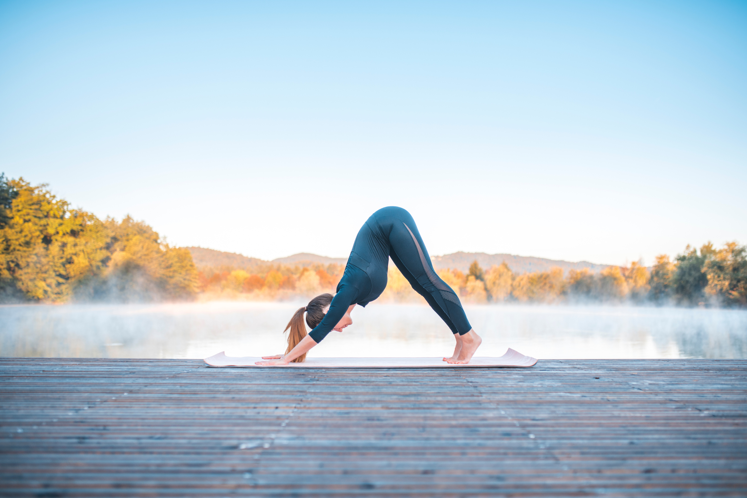 blonde woman with ponytail in all black on a wooden dock by water doing downward-facing dog yoga pose